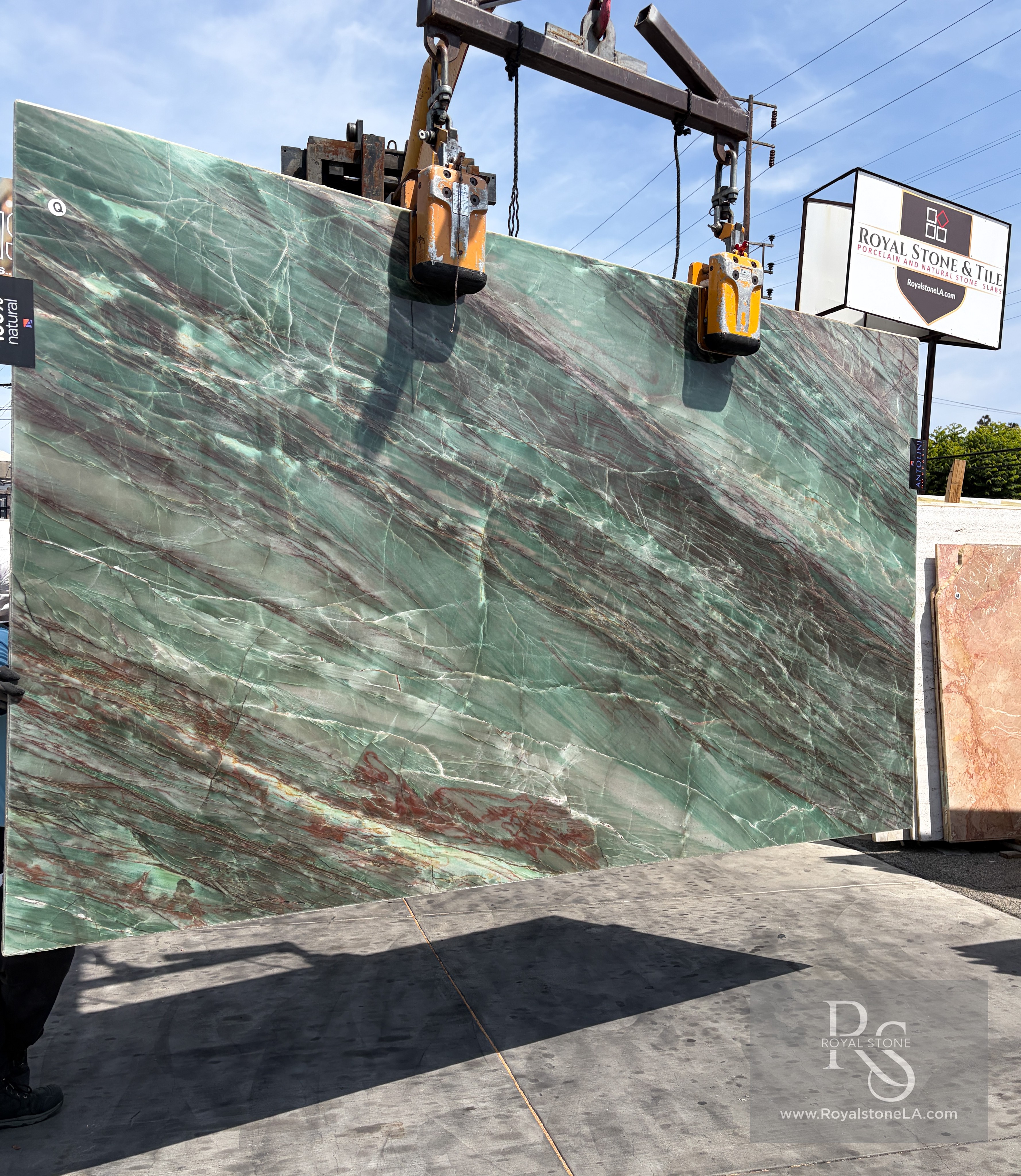 Antolini Thunderflash quartzite slab, 2cm polished, 76 by 129 inches, with pale seafoam-green field cut by diagonal garnet and copper veining, hanging from a crane at the Royal Stone & Tile Los Angeles yard