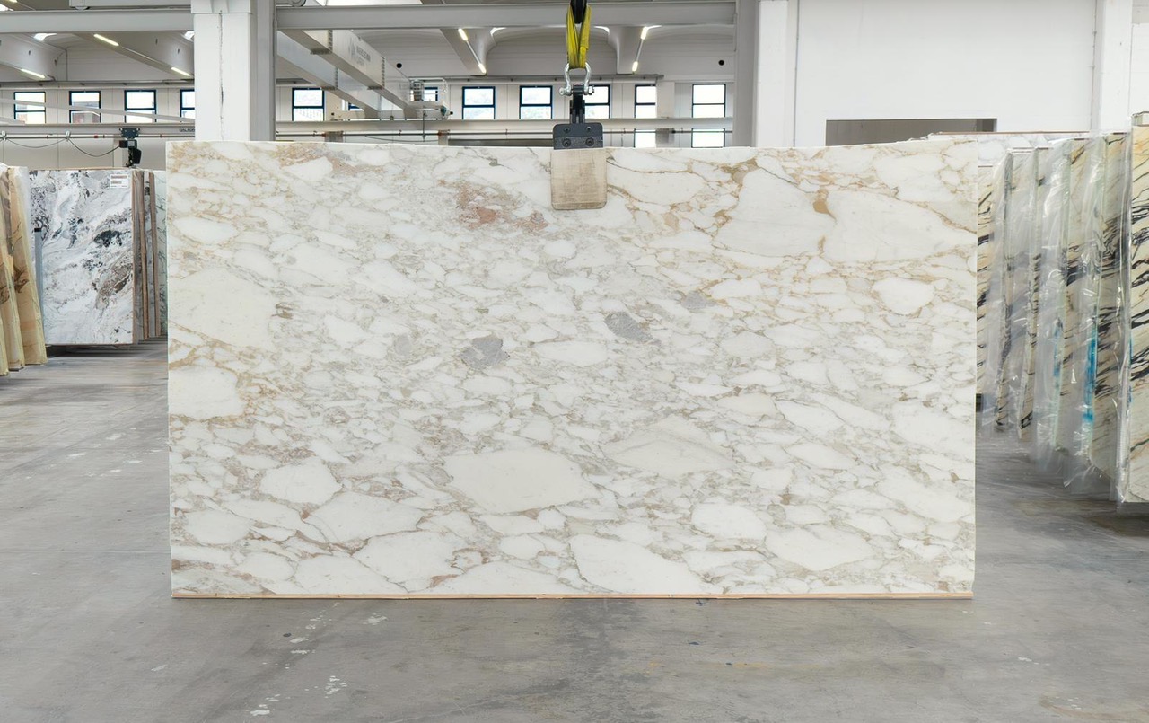 Italian Calacatta marble slab with white field and warm gold currents through soft grey veining, photographed at the factory