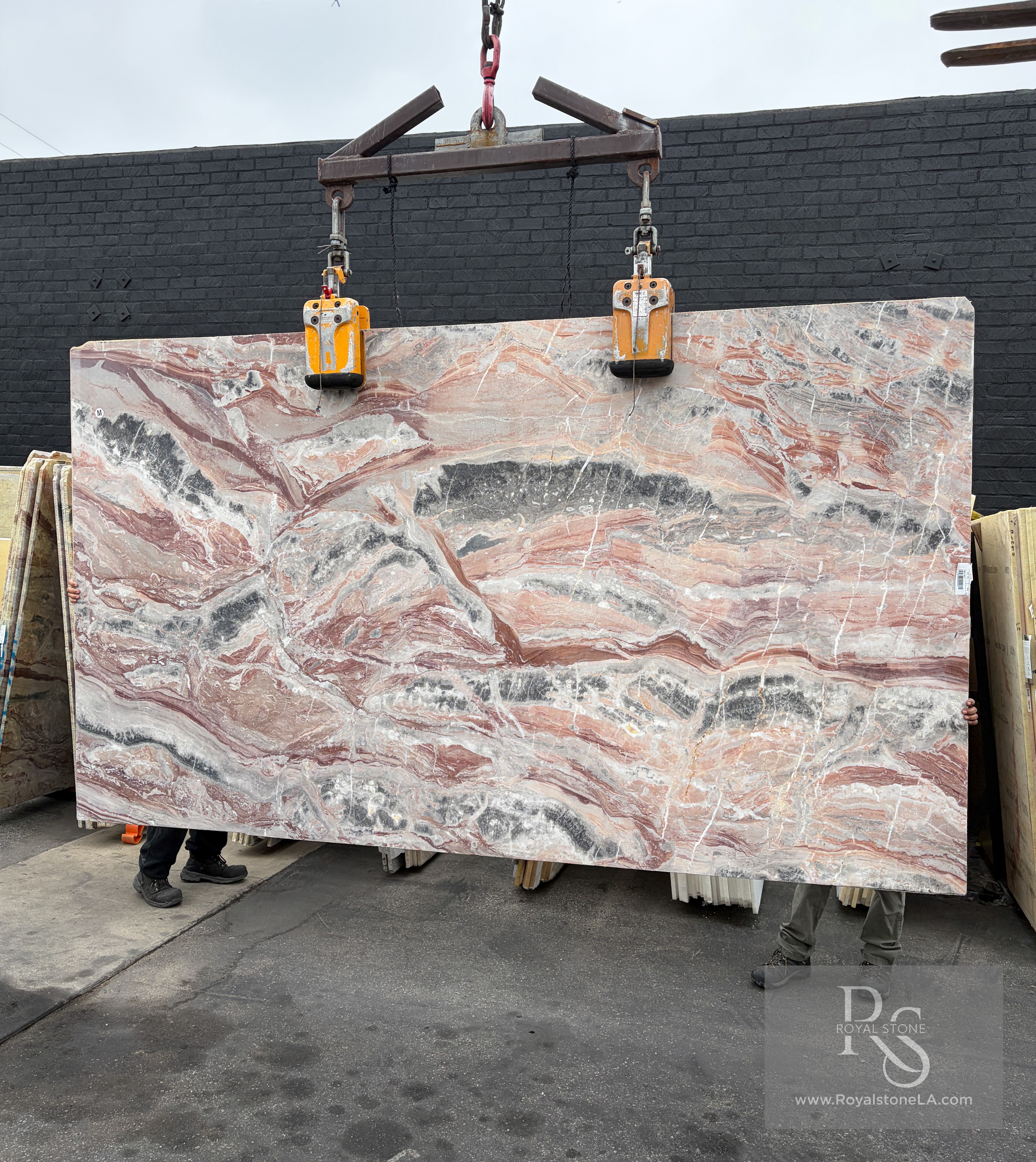 Viola Arabescato Orobico Rosso honed Italian marble slab, 75 by 130 inches, with horizontal rose, coral, charcoal, and white quartz currents, suspended from a crane in front of the Royal Stone & Tile yard wall